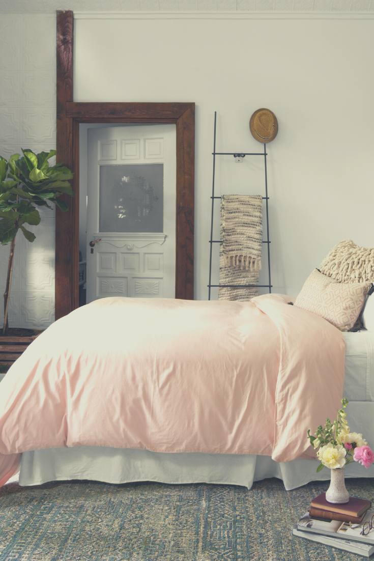 A queen bed with a pink blanket is shown, underneath it is a grey and blue area rug.
